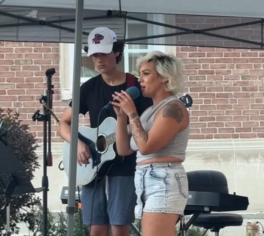 Miss Olivia performing outdoors with a student, singing and playing acoustic guitar